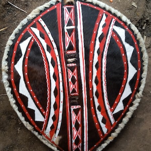 May include: A traditional shield made of animal hide with a black and white geometric pattern and red accents. The shield is decorated with a fringe of white fur.