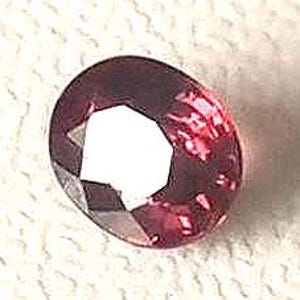 Puede incluir: Una gema facetada, de corte ovalado, con un profundo tono púrpura rojizo. La superficie de la gema refleja la luz, mostrando sus múltiples facetas y estructura interna. La piedra se encuentra sobre un fondo blanco texturizado.