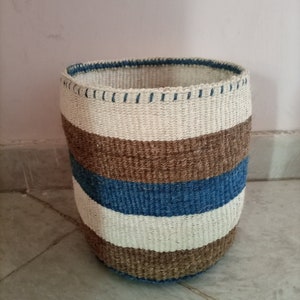 Puede incluir: Una cesta de almacenamiento tejida con rayas marrones, blancas y azules. La cesta está hecha de fibras naturales y tiene una forma redonda.