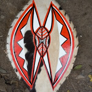 May include: A shield with a natural fur backing and a central design in red, white, and black. The shield's shape is oval, with a central design featuring geometric shapes and patterns. The fur has patches of black, brown, and white.