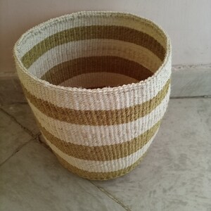 Könnte beinhalten: Ein geflochtener Aufbewahrungskorb mit einem gestreiften Muster in Beige und Braun. Der Korb ist aus Naturfasern gefertigt und hat eine zylindrische Form.