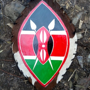 May include: A shield with a painted design featuring the colors of the Kenyan flag: black, red, green, and white. The shield is bordered by brown animal hide with white and brown fur.