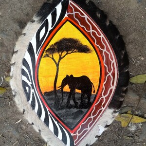 May include: A hand-painted shield with a zebra pattern on the left side and a red and white pattern on the right side. The center of the shield features a yellow sunset with a silhouette of an elephant and a tree.