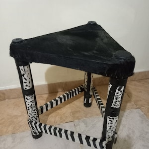 Puede incluir: Taburete de tres patas con asiento triangular cubierto de piel negra. Las patas están pintadas en blanco y negro con un patrón de cebra. El taburete mide aproximadamente 46 cm de altura.