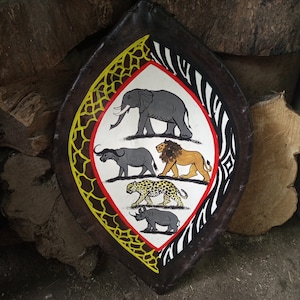 May include: A round, black leather shield with a white center featuring a colorful illustration of five African animals: an elephant, a lion, a leopard, a rhinoceros, and a zebra. The shield is decorated with a yellow and black pattern.