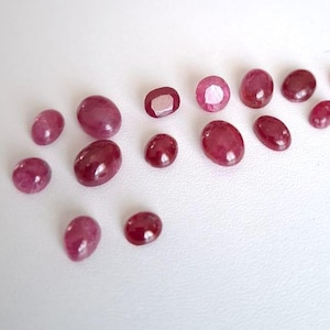 Könnte beinhalten: Eine Sammlung von 14 ovalen Rubin-Edelsteinen in verschiedenen Größen, die auf einer weißen Oberfläche präsentiert werden.