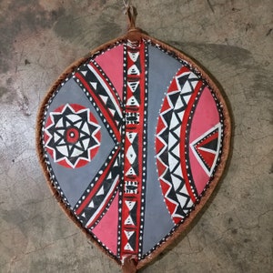 May include: A gray and pink shield with a geometric pattern. The shield is decorated with black and white lines and shapes. The shield is made of leather and has a fringe around the edge.