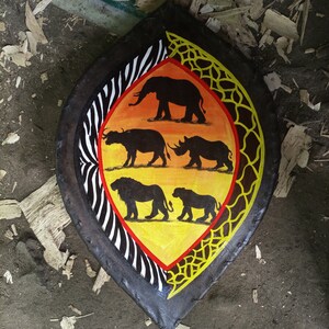 May include: A wooden shield painted with a yellow, orange, and black zebra pattern. The shield also features a black silhouette of an elephant, a rhino, a buffalo, and two lions.