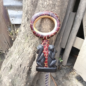 Puede incluir: Un bastón de madera con un patrón de jirafa tallado y dos monos en la parte inferior. El bastón está pintado en tonos de rojo, marrón y negro.