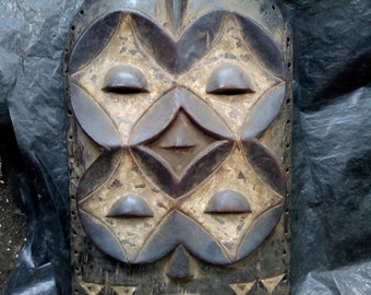 May include: A dark brown, carved wooden mask with a rectangular shape and rounded top. The mask features symmetrical, geometric designs, including four diamond-shaped elements and a central triangle. It has eye and mouth openings, and a handle at the top.