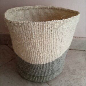 Könnte beinhalten: Ein geflochtener Aufbewahrungskorb mit einem weißen Oberteil und einem grauen Boden. Der Korb ist aus Naturfasern gefertigt und hat eine zylindrische Form.