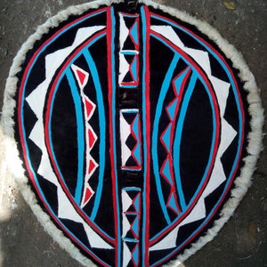 May include: A round shield with a black background and geometric patterns in white, red, and blue. The shield is made of leather and has a fringe around the edge.