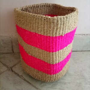 May include: A woven basket with pink stripes. The basket is made of natural fibres and has a cylindrical shape.