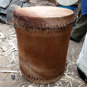 May include: A brown leather drum with a stitched seam around the top. The drum is sitting on a dirt floor with wood shavings around it.