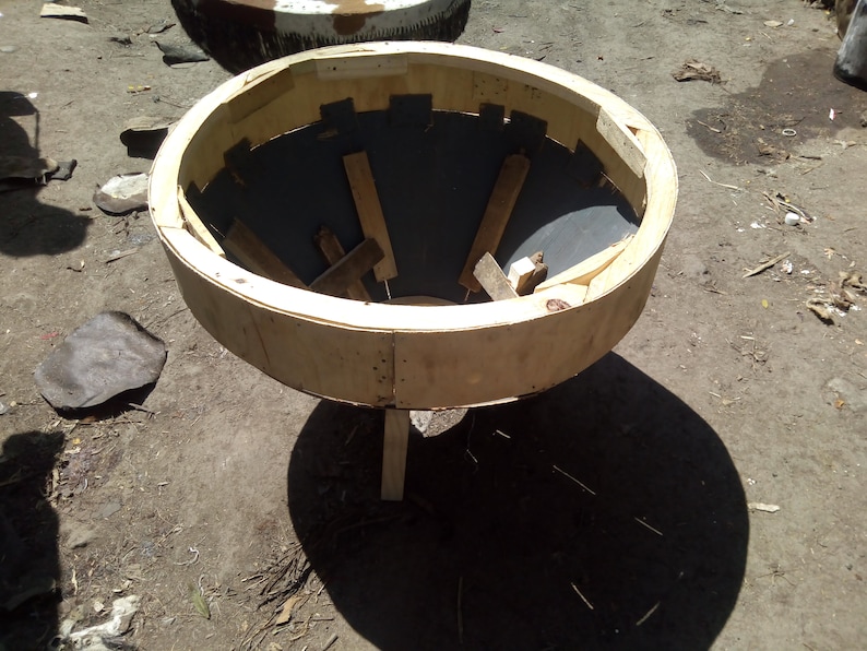 Puede incluir: Un molde de madera para hacer macetas de hormig&oacute;n. El molde est&aacute; hecho de madera contrachapada y tiene una parte superior circular con un fondo en forma de embudo. El molde est&aacute; sobre una superficie de tierra.