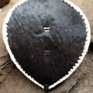 May include: A round, black leather shield with a white leather trim. The shield has two metal rivets on each side.