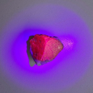 Puede incluir: Una gema de rubí rosa brillante, de corte tosco, bajo una luz UV, que emite un brillo púrpura.