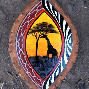 May include: A wooden shield with a painted design of a giraffe and a tree in a sunset landscape. The shield is decorated with a zebra pattern and red and white geometric designs.