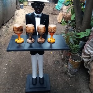 May include: A wooden figurine of a waiter wearing a white hat and a black tuxedo, holding a tray with four wooden cups. The figurine is standing on a black base.