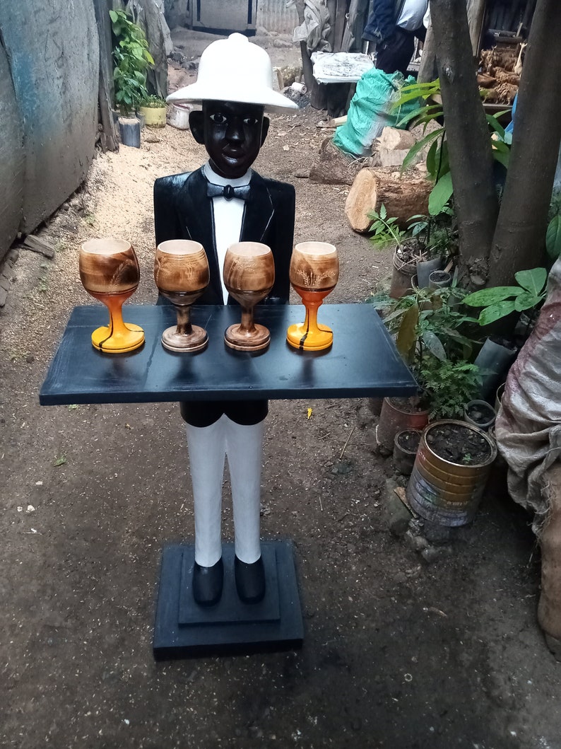 May include: A wooden figurine of a waiter wearing a white hat and a black tuxedo, holding a tray with four wooden cups. The figurine is standing on a black base.