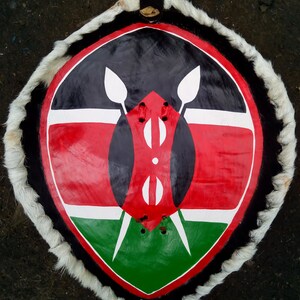 Könnte beinhalten: Ein runder Schild mit einem weißen Pelzbesatz. Der Schild ist in den Farben der kenianischen Flagge bemalt: Schwarz, Rot, Weiß und Grün. Der Schild ist ein traditionelles afrikanisches Design.