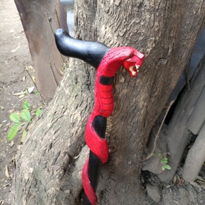 Puede incluir: Un bastón de caminar negro y rojo tallado en forma de serpiente. La serpiente está pintada de rojo con detalles negros. El bastón se apoya en un tronco de árbol.