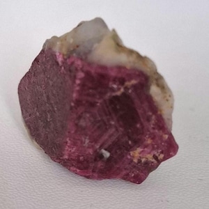 Puede incluir: Un cristal de rubí rojo intenso con una superficie rugosa y natural. El cristal está rodeado de matriz blanca y beige.