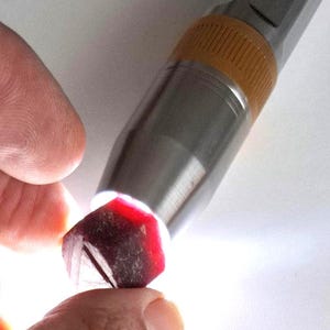 Pode incluir: Uma gema facetada, vermelho escuro, é segurada entre os dedos, iluminada por uma lanterna prateada. As bordas da gema são bem definidas e a luz revela sua estrutura interna. A imagem destaca a cor e a clareza da gema.