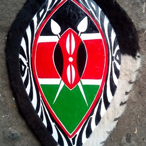 May include: A traditional shield with a black, white, and red zebra pattern. The center of the shield features the flag of Kenya, with a red, black, and green design. The shield is made of leather and has a fringe around the edge.