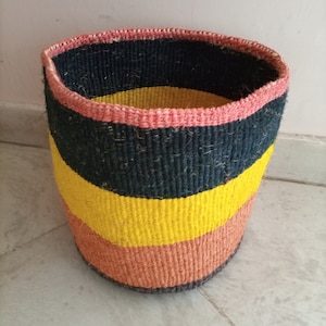 Könnte beinhalten: Ein geflochtener Aufbewahrungskorb mit einem schwarz-gelb-orangen Streifenmuster. Der Korb hat einen rosa Rand und ist aus Naturfasern gefertigt.
