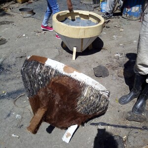 Puede incluir: Un tambor de piel de vaca marr&oacute;n y blanco con flecos negros, parcialmente ensamblado sobre una base de madera. El tambor est&aacute; sentado sobre una superficie de tierra junto a un marco de madera con un interior gris.