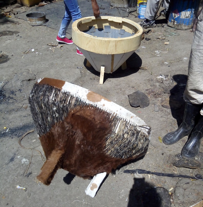 Puede incluir: Un tambor de piel de vaca marr&oacute;n y blanco con flecos negros, parcialmente ensamblado sobre una base de madera. El tambor est&aacute; sentado sobre una superficie de tierra junto a un marco de madera con un interior gris.