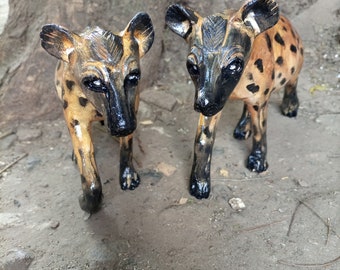 Puede incluir: Dos pequeñas figuritas de perros salvajes africanos pintadas a mano. Las figuritas están hechas de un material marrón claro y tienen marcas negras. Están de pie sobre una superficie marrón, similar a la tierra.