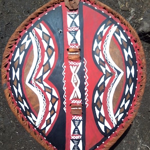 May include: A traditional African shield with a red, black, and white geometric pattern. The shield is made of leather and has a fringe around the edge.