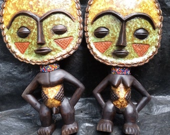 May include: Two wooden sculptures of stylized human figures with circular faces. The faces are painted in shades of green, orange, and brown with a gold-like finish. The figures are wearing beaded necklaces and have a gold-like finish on their torsos.