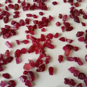 Op de afbeelding: Een verzameling losse rode robijn edelstenen in verschillende maten en vormen. De robijnen zijn verspreid over een wit oppervlak.