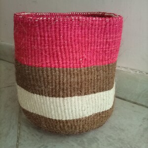 May include: A woven storage basket with a pink, brown, and white striped pattern. The basket is made of natural fibers and has a cylindrical shape.
