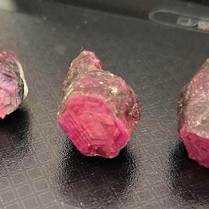 May include: Three rough-cut ruby gemstones with a deep red hue. The stones have a crystalline structure and are partially encased in a darker, possibly weathered, matrix. The gems are displayed on a dark surface.