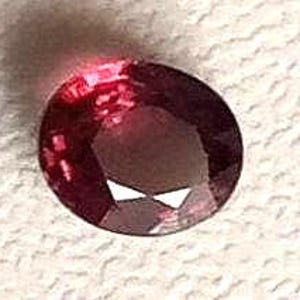 May include: A deep red, oval-cut gemstone with multiple facets. The gem's cut creates a sparkling effect, reflecting light. The stone is set against a textured, off-white surface.