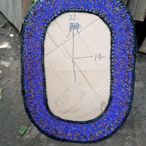 May include: A blue and black beaded oval mirror frame with a cardboard backing. The cardboard has handwritten measurements of 21 inches by 19 inches.