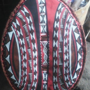 May include: A round shield with a red, black, and white geometric pattern. The shield is made of fabric and has a fringe around the edge.