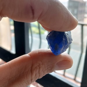 Op de afbeelding: Een diepblauwe edelsteen met een ruw, gefacetteerd oppervlak. De edelsteen wordt tussen twee vingers gehouden.