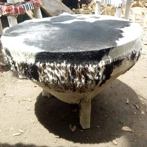 Puede incluir: Un tambor redondo de piel de vaca negra y blanca con una base de madera y tres patas. El tambor est&aacute; hecho de materiales naturales y tiene un aspecto r&uacute;stico.