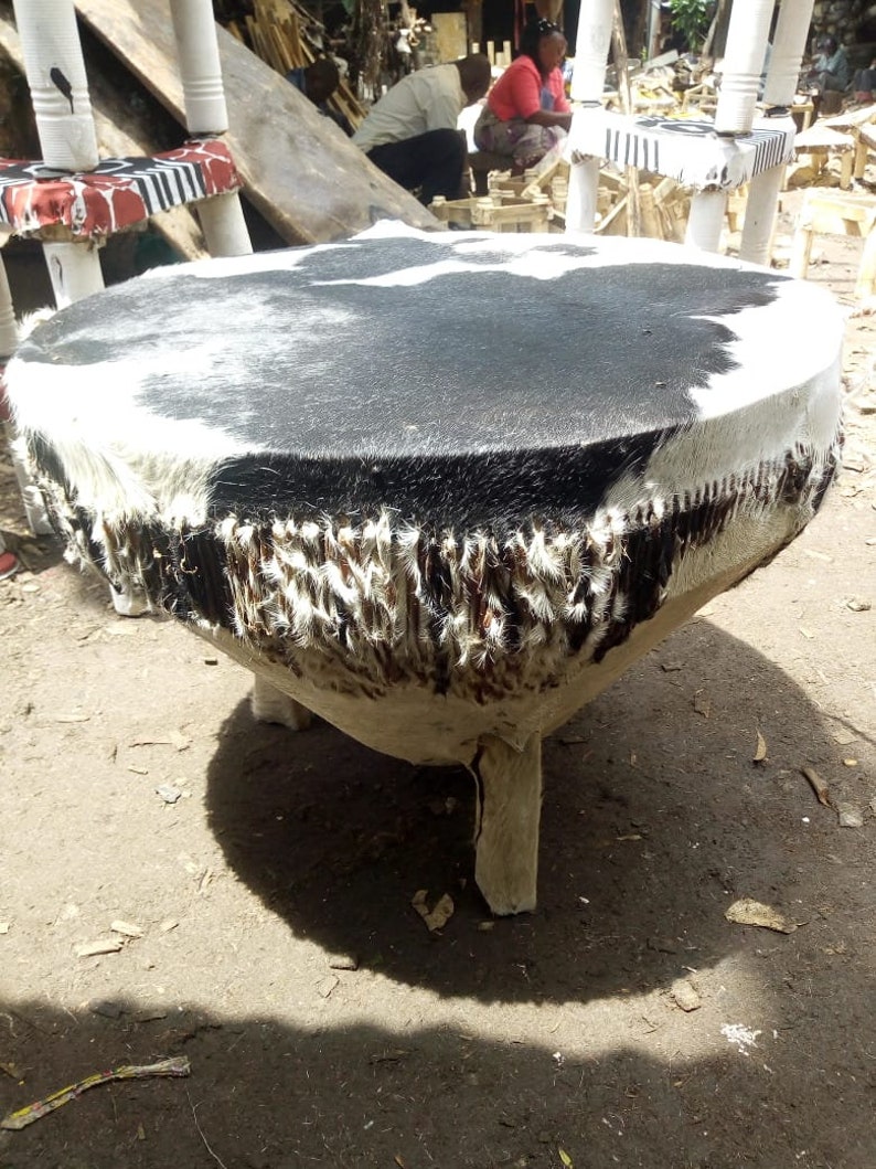 Puede incluir: Un tambor redondo de piel de vaca negra y blanca con una base de madera y tres patas. El tambor est&aacute; hecho de materiales naturales y tiene un aspecto r&uacute;stico.