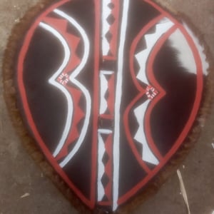 Könnte beinhalten: Ein traditioneller Schild mit einer braunen Lederbasis und einem geometrischen Muster, das in Rot, Weiß und Schwarz bemalt ist. Der Schild ist oval geformt und hat einen Fransen aus braunem Fell am Rand.