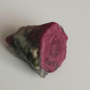 Peut inclure: Un cristal de rubis rose avec une forme hexagonale et une texture brute et naturelle. Le cristal est entouré d'une matrice grise et blanche.