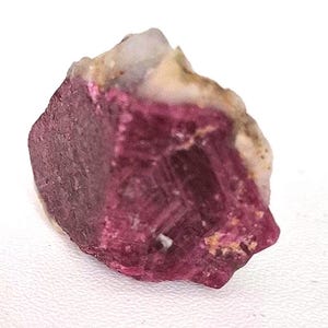 Pode incluir: Uma gema bruta, não cortada, de um magenta profundo, com uma estrutura cristalina. A pedra tem uma superfície rugosa e texturizada com áreas de depósitos minerais brancos e bege. A forma é irregular, mostrando formações geológicas naturais.