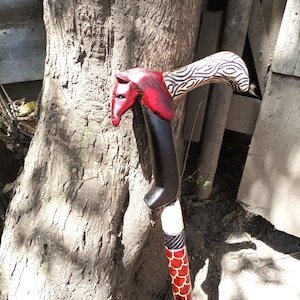 Puede incluir: Un bastón de madera con un mango tallado en forma de cabeza de caballo. El mango está pintado de rojo y negro, y el eje está pintado con un patrón blanco y negro.