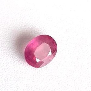 Könnte beinhalten: Ovale geschliffener Rubin-Edelstein mit facettierter Oberfläche. Der Edelstein hat eine leuchtende, rötlich-rosa Farbe und liegt auf einer weißen Oberfläche. Nahaufnahme des Edelsteins, die seine Klarheit und Brillanz hervorhebt.