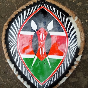 May include: A round shield with a black, white, red, and green design. The shield is made of leather and has a zebra print border. The center of the shield features the flag of Kenya.